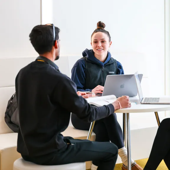 Two female students sit at a round table talking to a male student who has his back turned to us. They have laptops in front of them