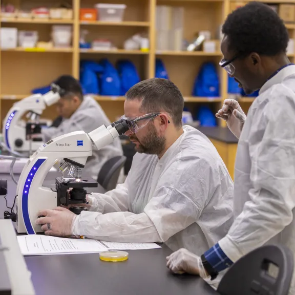 A couple students look through microscopes in a lab while an instructor looks on