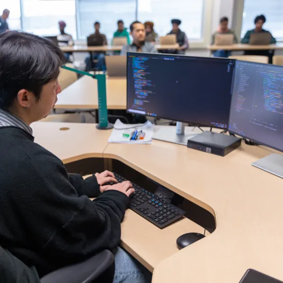 A male student sits coding in front of two screens in a classroom full of fellow students