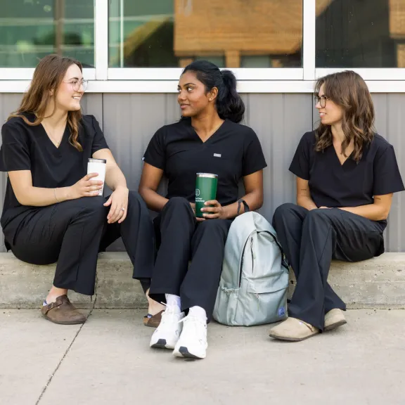 Three healthcare students sit outside chatting, in their RDP black scrubs with RDP branded coffee mugs.