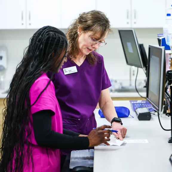 An instructor in purples scrubs works with a student in pink scrubs at a pharmacy desk.