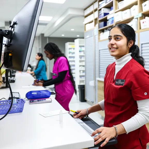 A student in red scrubs looks at a computer in a pharmacy.