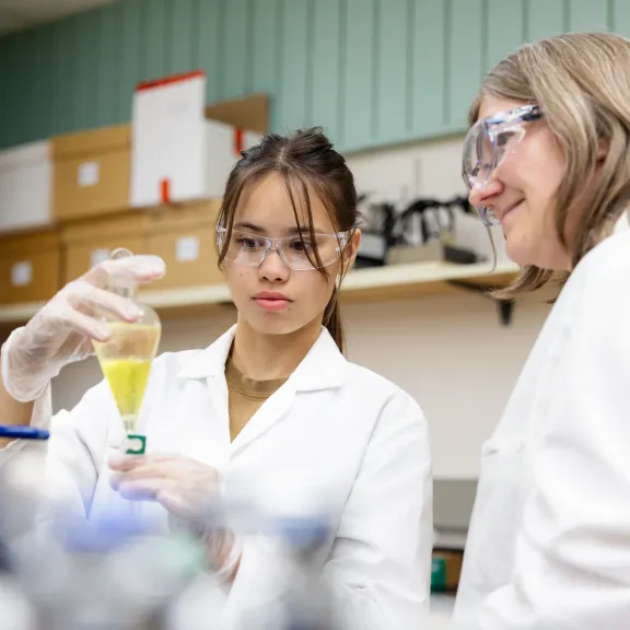 A student and instructor in a chemistry lab with a teardrop shaped beaker full of yellow liquid.