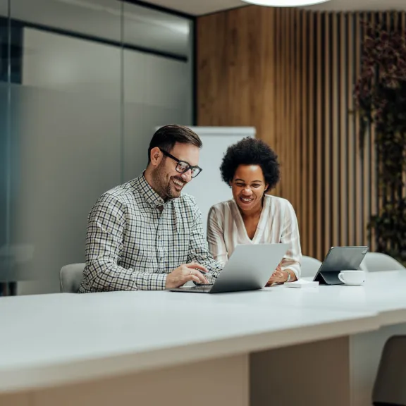 A man in glasses and plaid shirt and a woman in a grey top sit beside each other at a table smiling at a laptop in an office