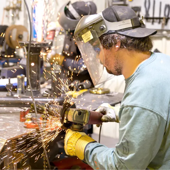 Man in protective gear grinds some metal in a shop as sparks fly