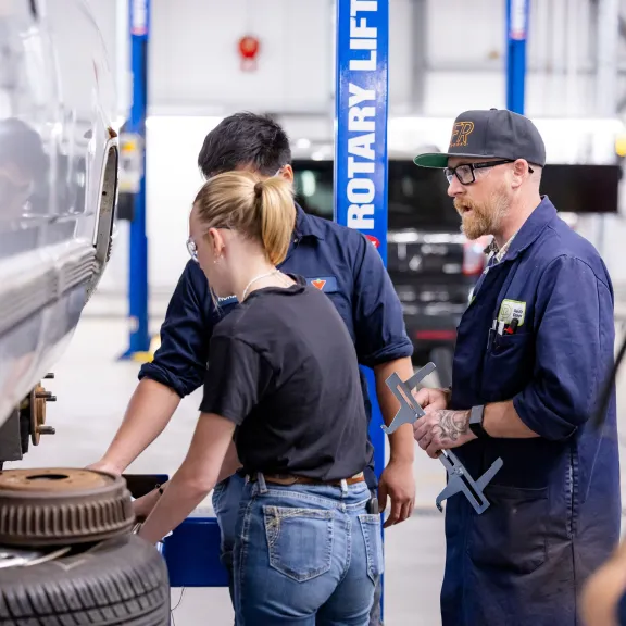 A male and female student and an instructor work on a truck in a shop