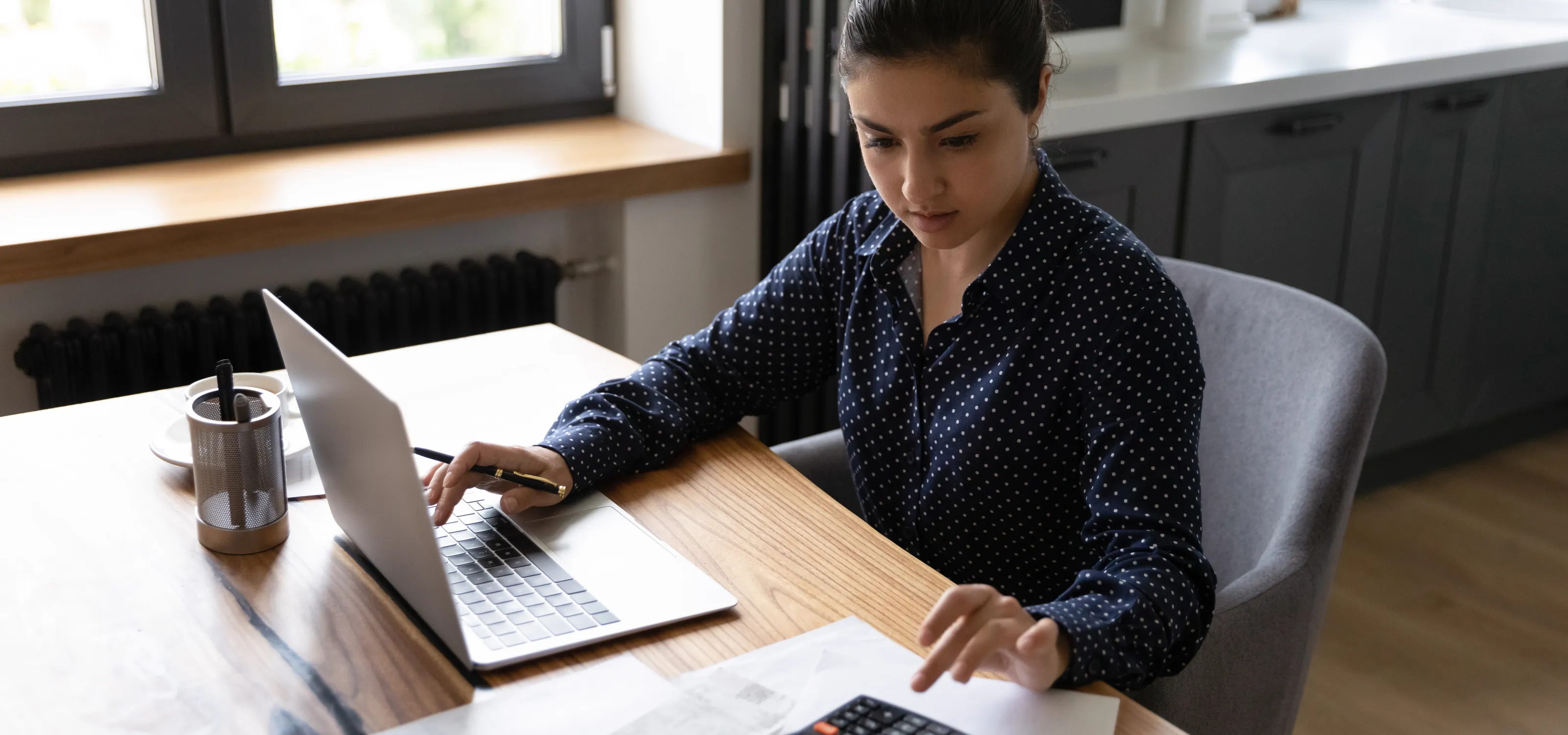 Young woman working on her laptop while entering digits on a calculator.