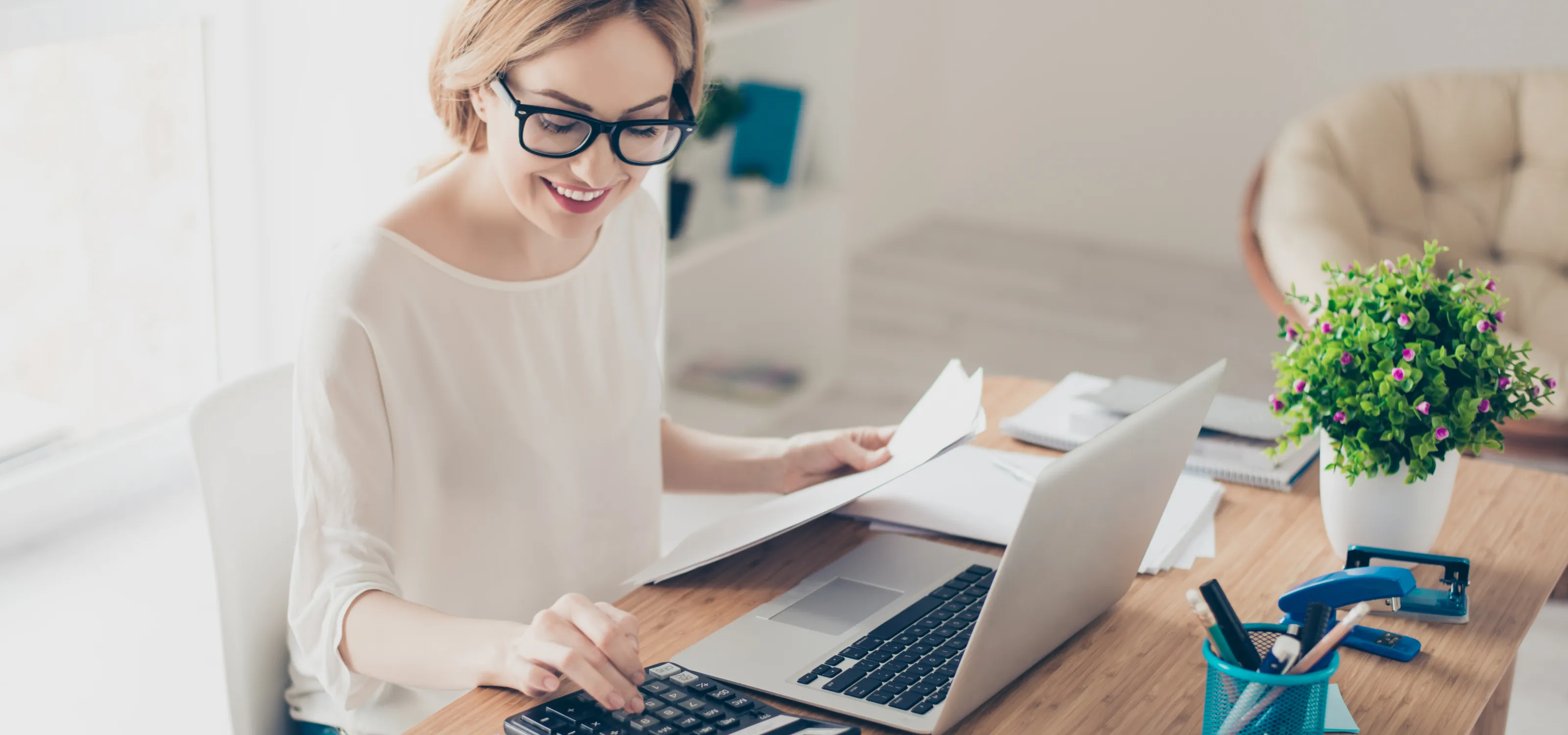 Woman working on a calculator holding papers with her laptop open.