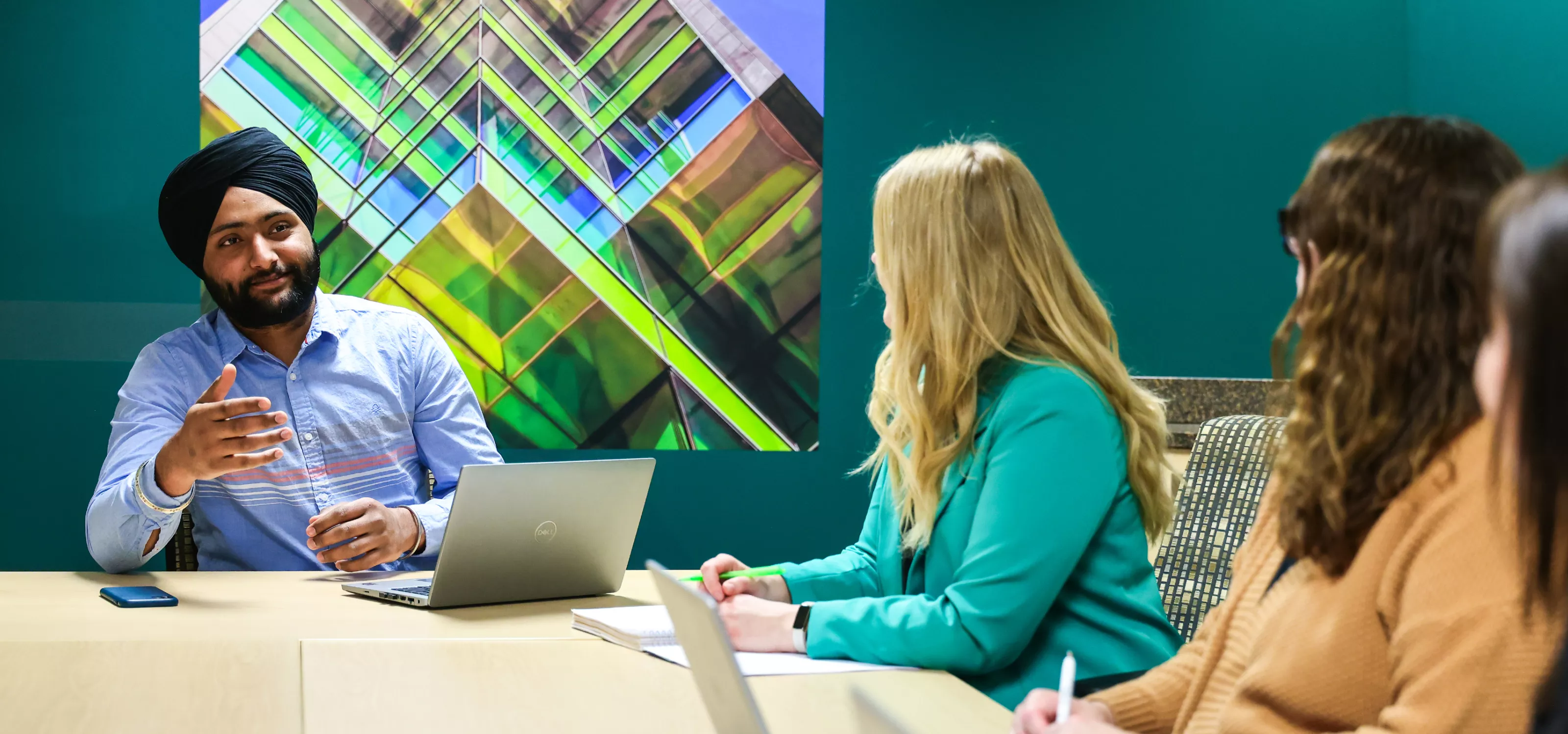 A group of professionals at a board table in a green room. On the wall hangs a large photo of a skyscraper with bright green segments dividing each floor. A man in a blue shirt and black headwrap smiles while talking to his coworkers whose faces are not shown to camera. 