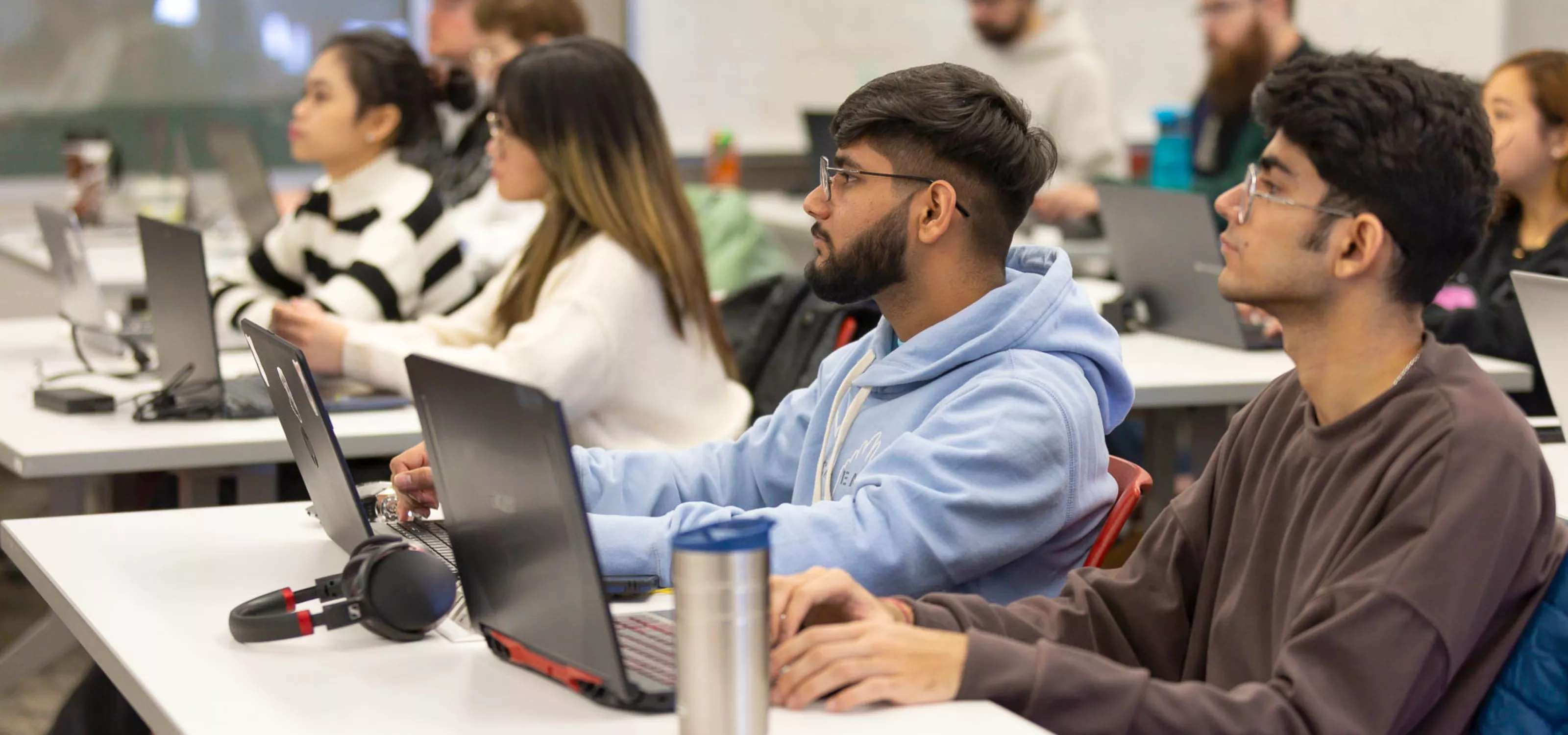 A diverse group of students sits on their laptops in a classroom looking towards a teacher who is off-screen.