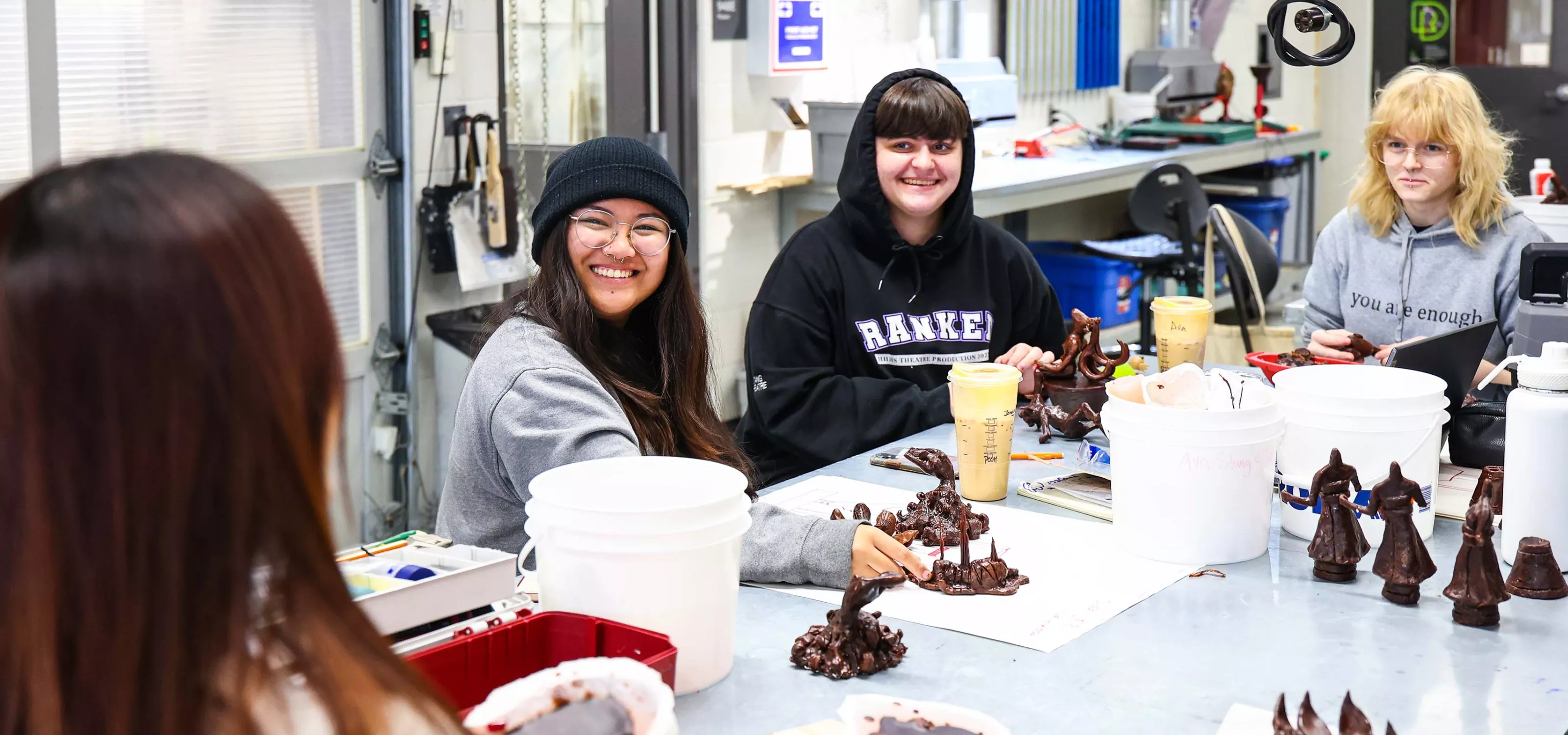 A group of smiling students sit in front of a table full of their sculpture creations