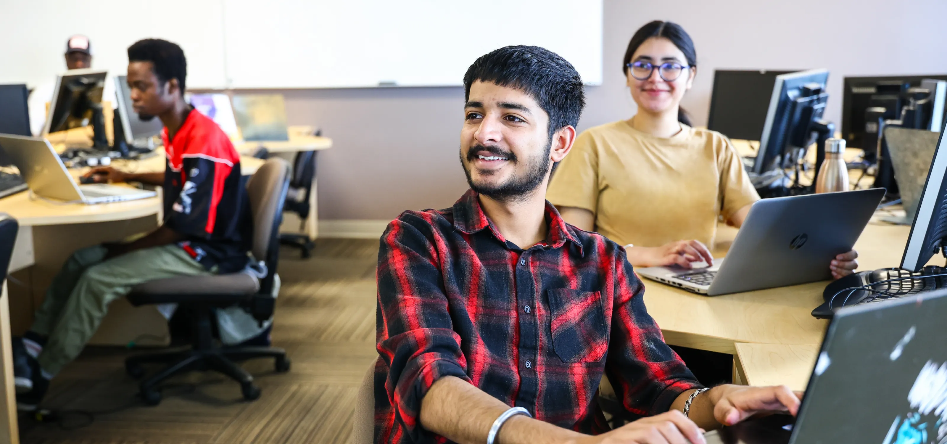 A, student in a plaid shirt, smiles to the instructor (who is not seen) in a computer lab classroom.