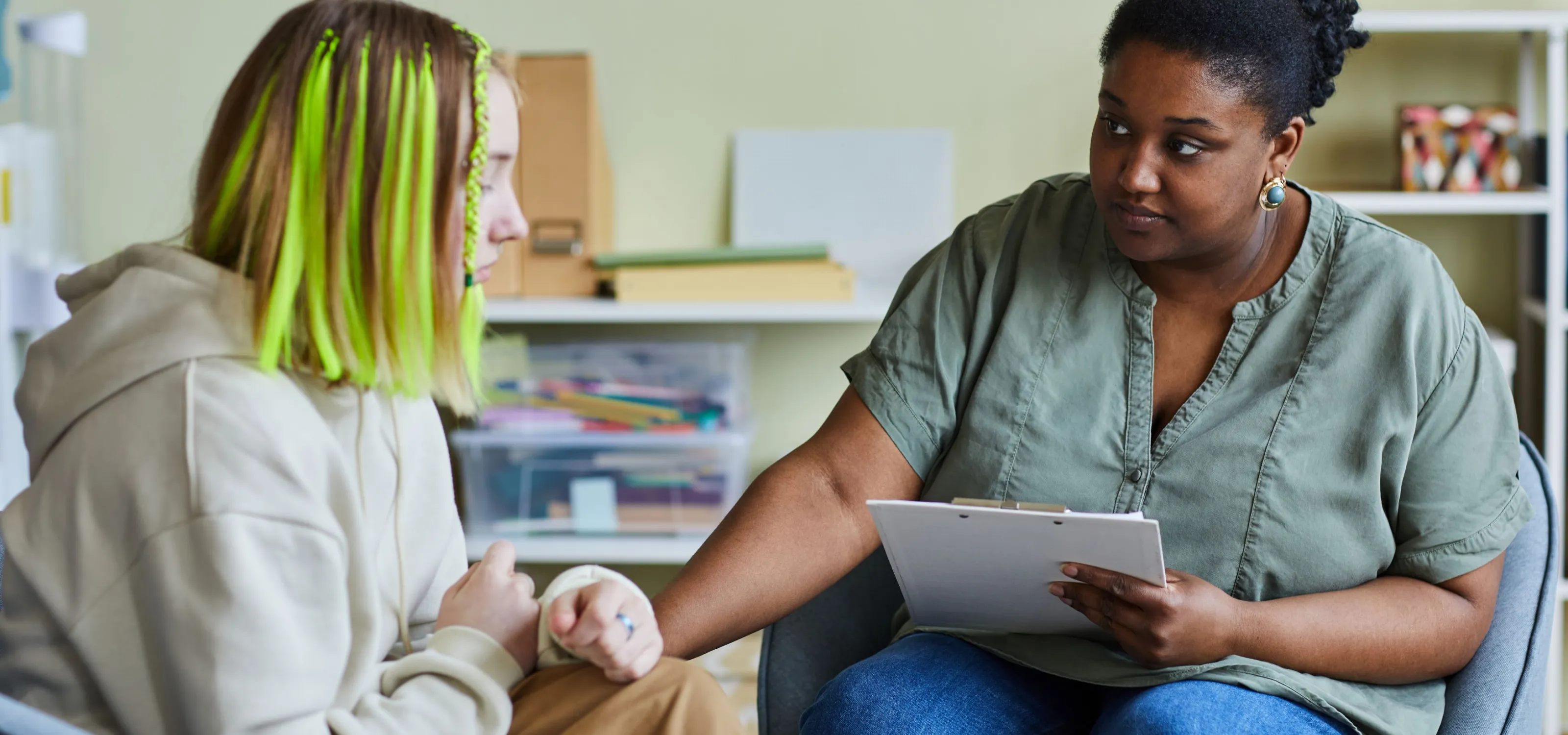 A Black woman in scrubs looks kindly and places a hand of support on a sad teenager