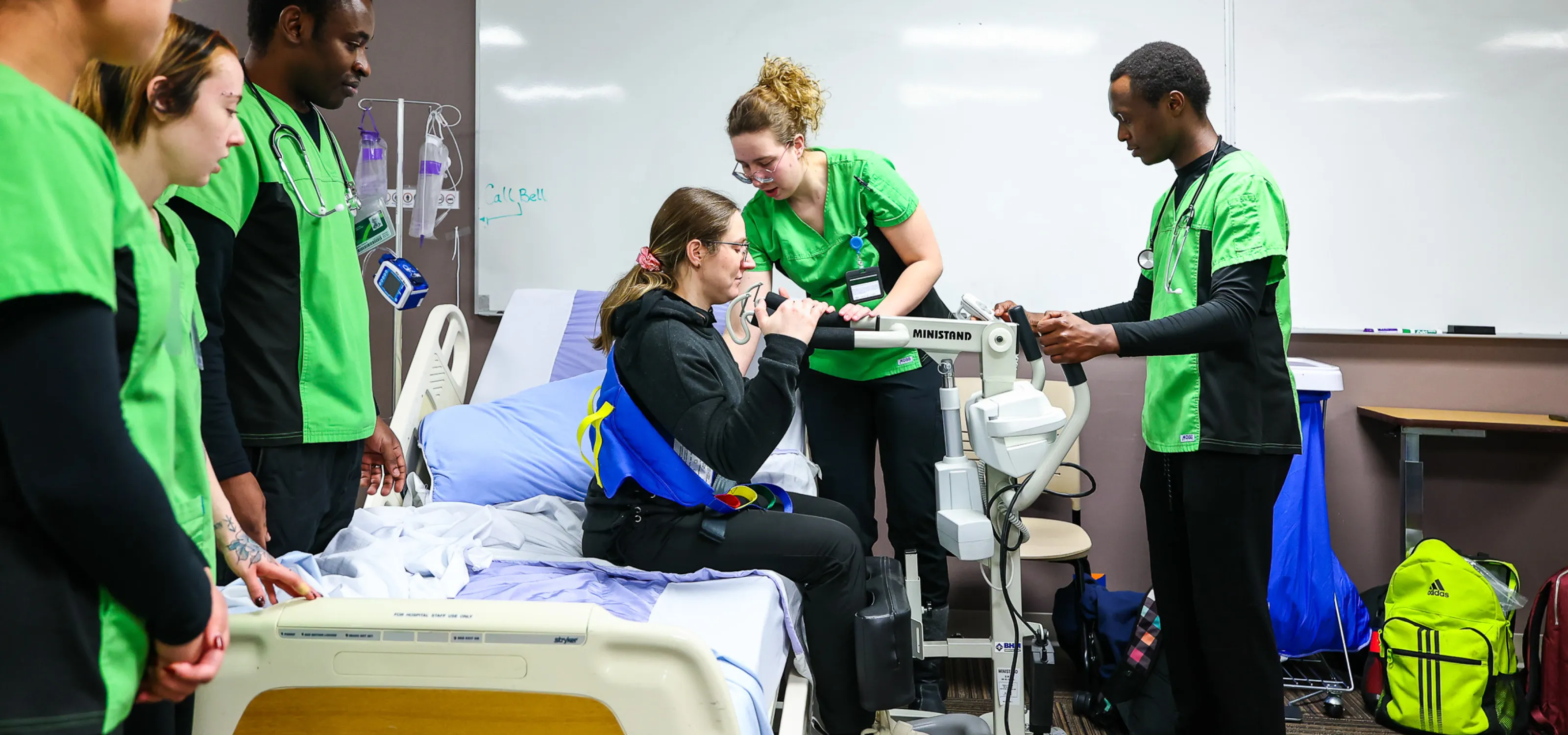 health care aide students, in lime green scrubs, help a patient stand up from a seated position using assistive device.