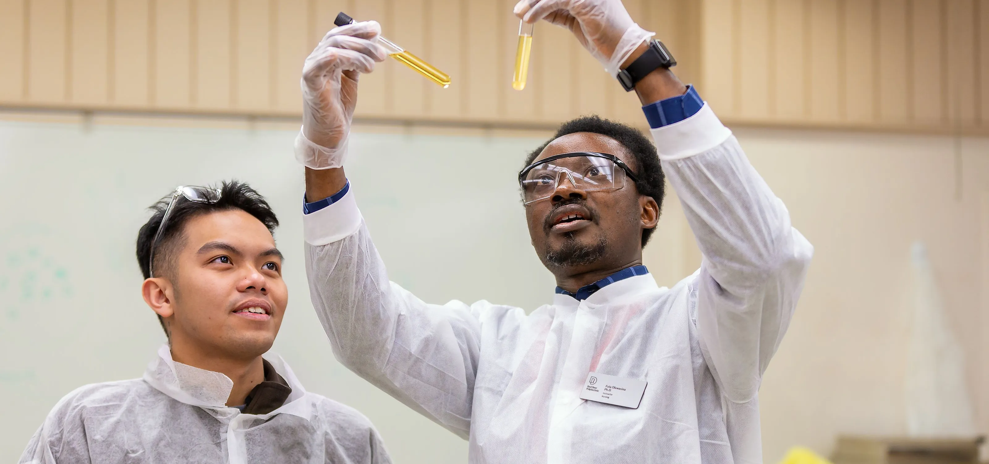 An instructor wearing full PPE holds up a couple of test tubes into the light and looks at them A student on his left looks on