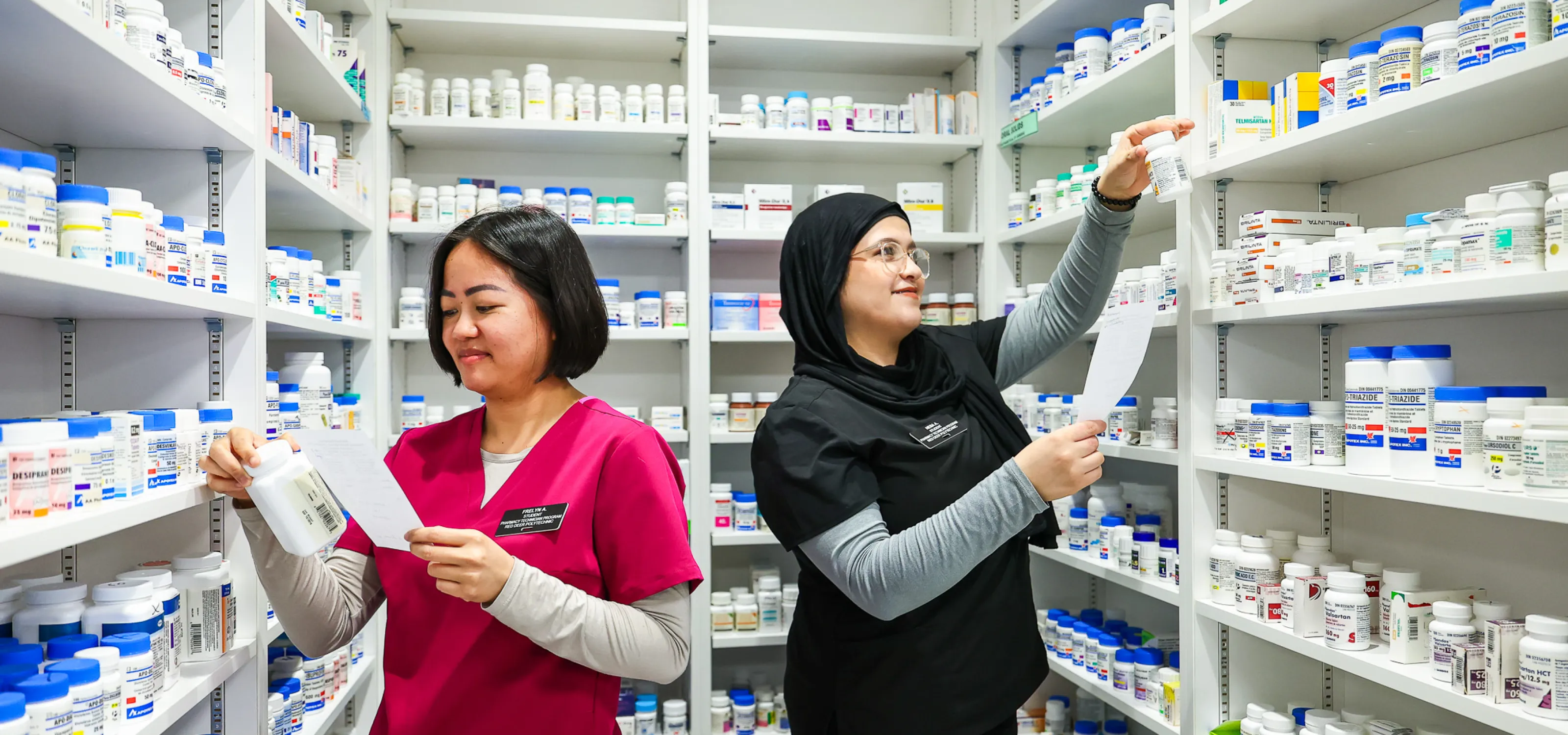Two pharmacy students look for medications in shelves on three walls around them.