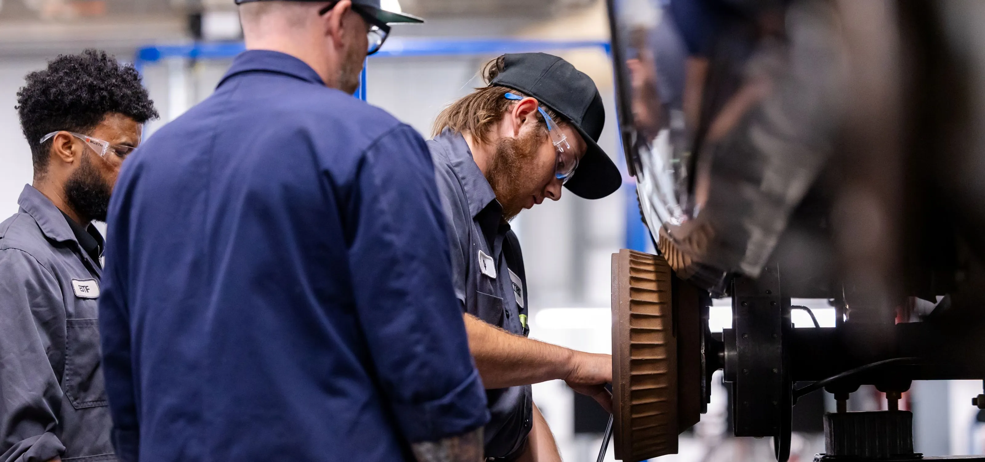 Two students and an instructor in blue coveralls work on a truck rim in a shop